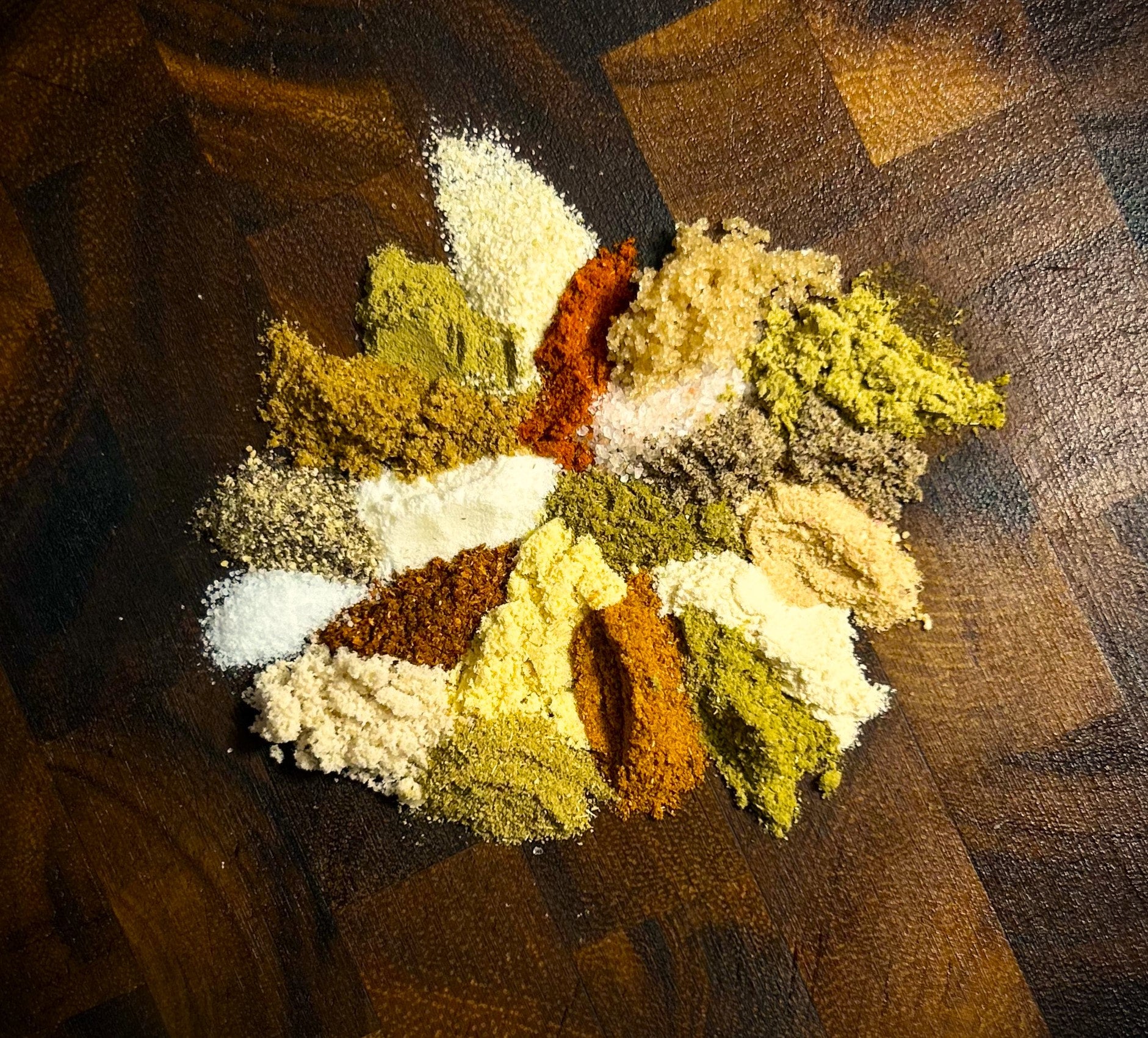 20 individual spices, used to craft Blue Ember Spice Rub, placed in a spiral on a deep brown cutting board